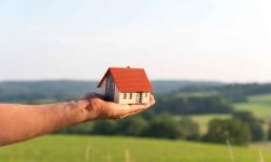 Auf dem Bild ist eine Hand zu sehen, die ein Miniaturhaus trägt. Im Hintergrund ist eine grüne Landschaft zu erkennen.