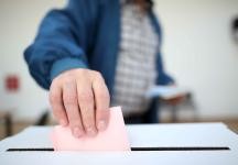 Hier ist ein Foto von einer Hand zu sehen, die einen rosa Wahlzettel in eine Wahlurne steckt. Die Hand gehört zu einer Person, die eine blaue Jacke und ein kariertes Hemd trägt. Der Hintergrund ist unscharf, wodurch der Fokus auf die Hand und die Wahlurne gelegt wird. 